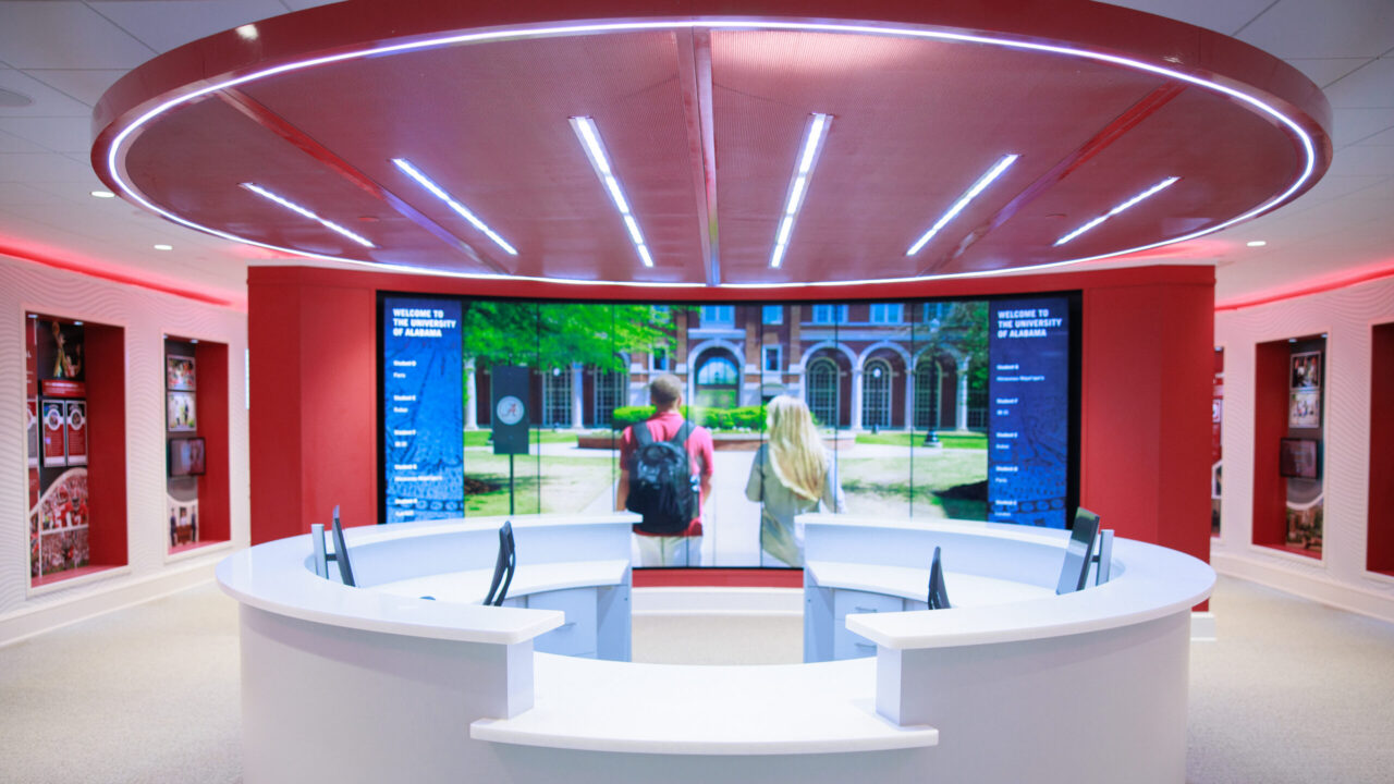 A futuristic welcome center at The University of Alabama featuring a circular information desk with a large digital display showing the university's campus. The room has a bright red theme with modern lighting and provides an interactive area for visitors.
