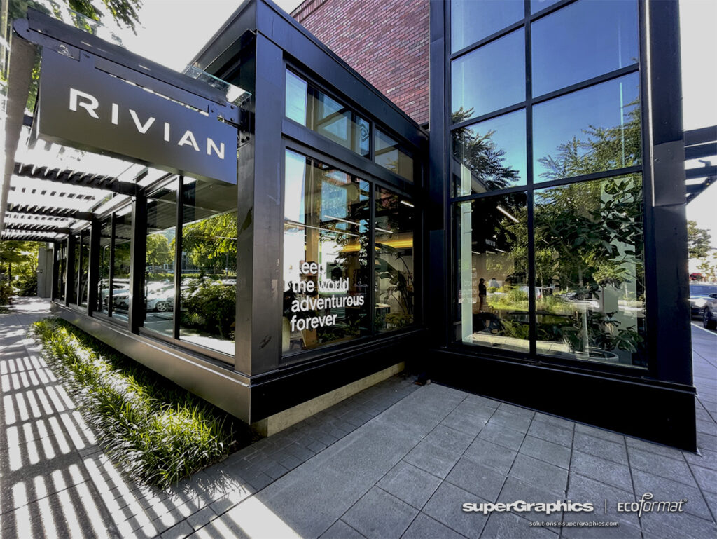 The exterior view of a Rivian showroom in Seattle, featuring prominent window graphics that promote the brand's adventurous spirit. The modern glass facade and the lush greenery surrounding the showroom create an inviting atmosphere.