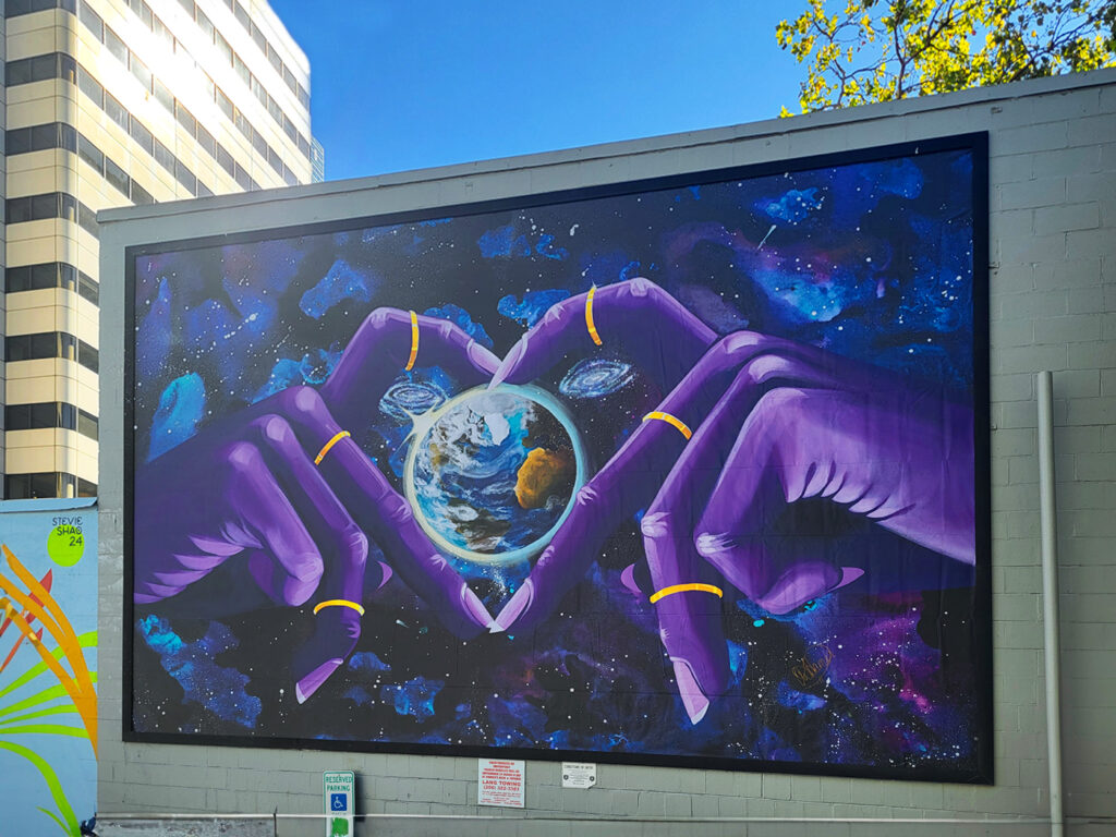 Vibrant mural of two large, purple hands forming a heart shape while holding Earth against a cosmic background, with galaxies and stars swirling around.