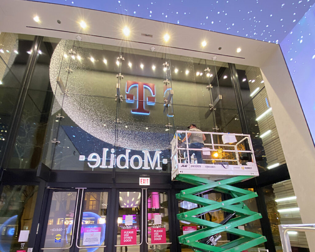 Graphic installation for T-Mobile Holiday, produced and installed by SuperGraphics. Showcases graphic installer applying white vinyl globe graphics on T-Mobile Storefront window.