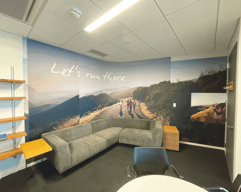Photo-realistic wall mural in a Brooks Running office lounge showing runners on a mountain trail with the text 'Let's run there' to reinforce brand ethos.