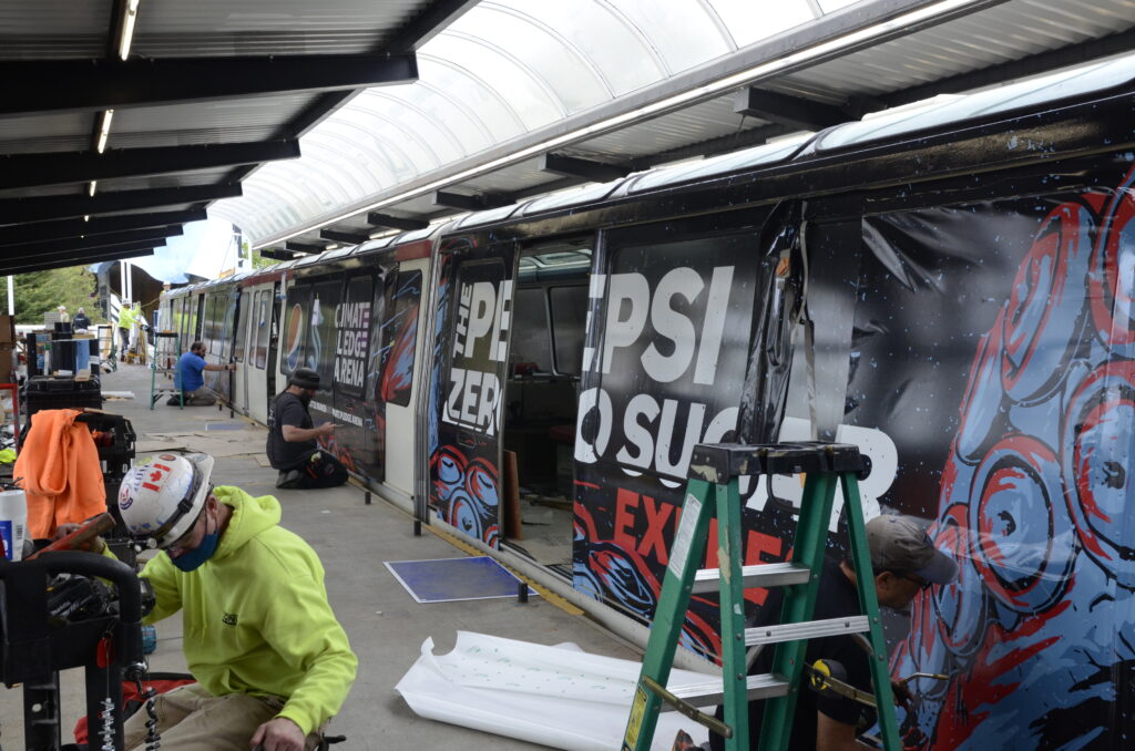 Installers applying large format vinyl transit wrap for the Seattle Kraken and Pepsi sponsorship campaign onto the side of a monorail car.