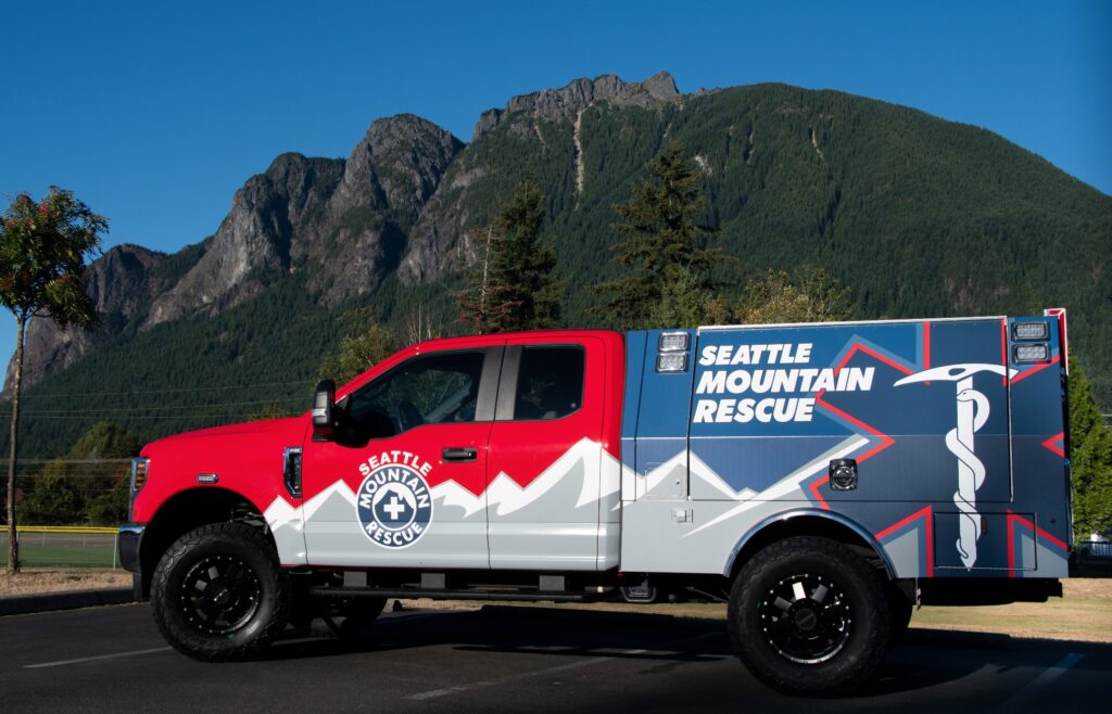 Seattle Mountain Rescue utility truck with a custom vinyl wrap featuring a mountain landscape design, the organization's logo, and red and blue colors.