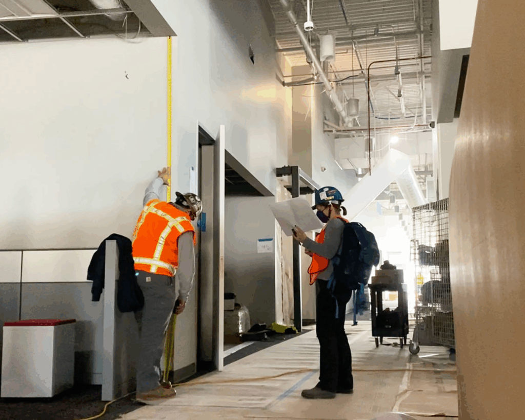 Photo of SuperGraphics team conducting site survey on a construction site at T-Mobile Kingsburg Call Center. Showcases one person in PPE measuring wall height, while another person looks at map of project scope.