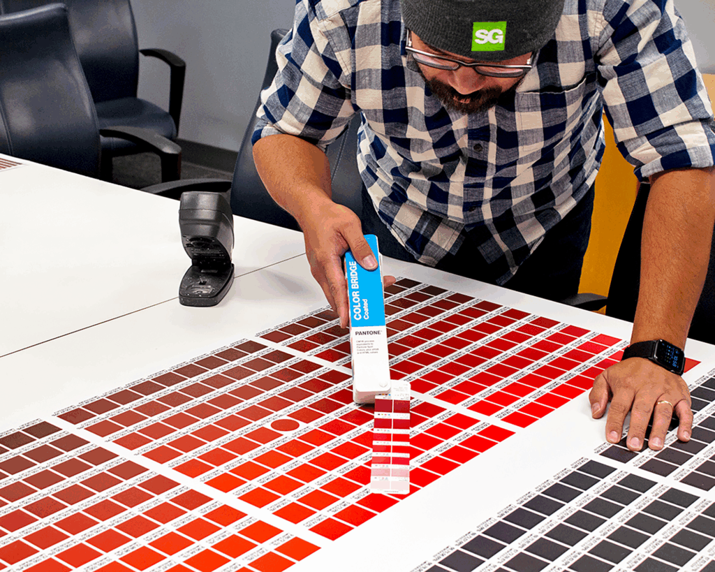 Graphics production expert using a color calibration tool on a large printed color swatch sheet to ensure brand-obsessed color precision.