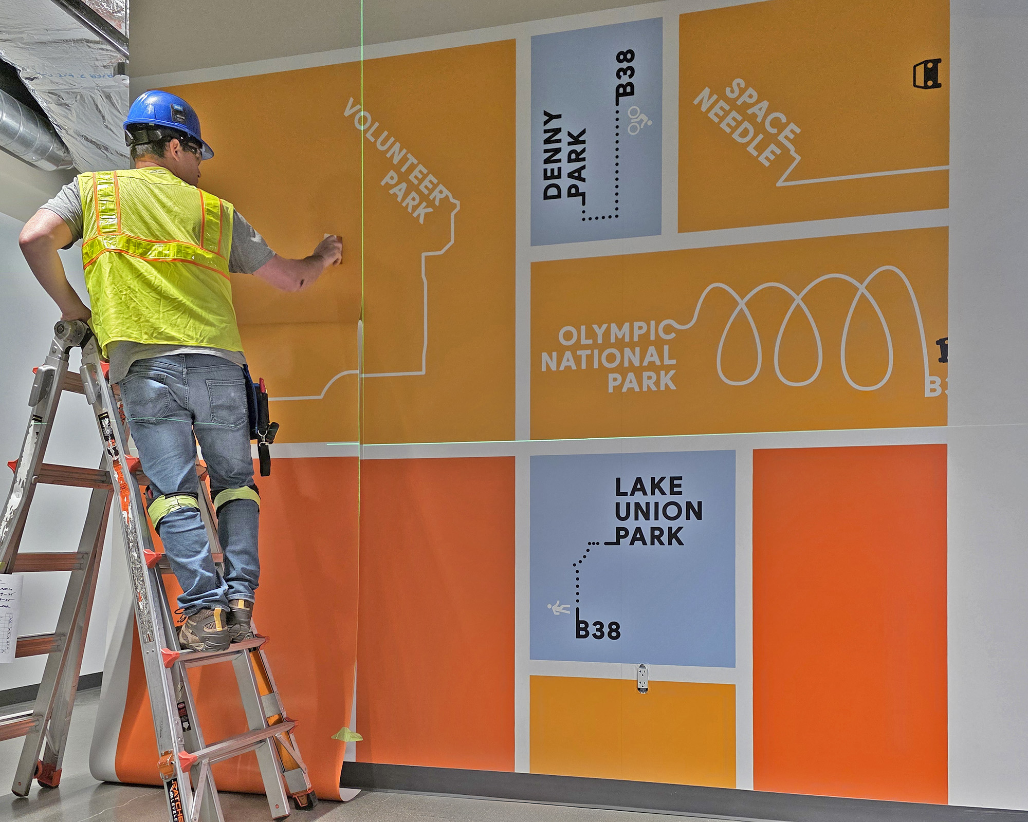 Photo of SuperGraphics installing wall mural at construction site. Features installer in PPE squeegeeing wall mural with Seattle and Washington State landmark graphics.
