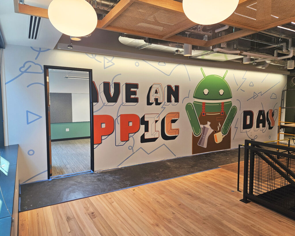 Photo of graphic mural installation completed on construction site, produced and installed by SuperGraphics. Wall mural graphic showcases "Have an Appic Day" with an Android icon pouring coffee.