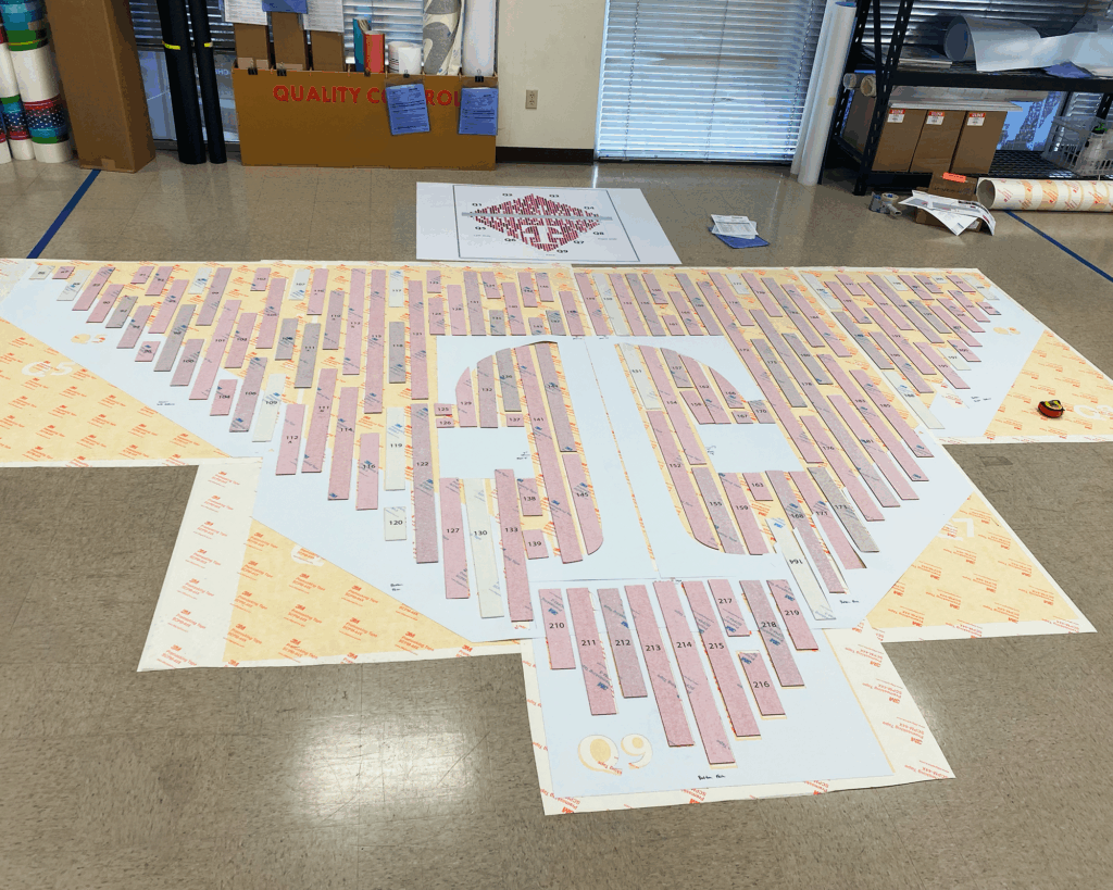 Photo of QC layout of acrylic panels for T-Mobile graphic installation, produced by SuperGraphics. Showcases pink acrylic pieces assembled to create T-Digit, all numbered out, under premasking protective paper.
