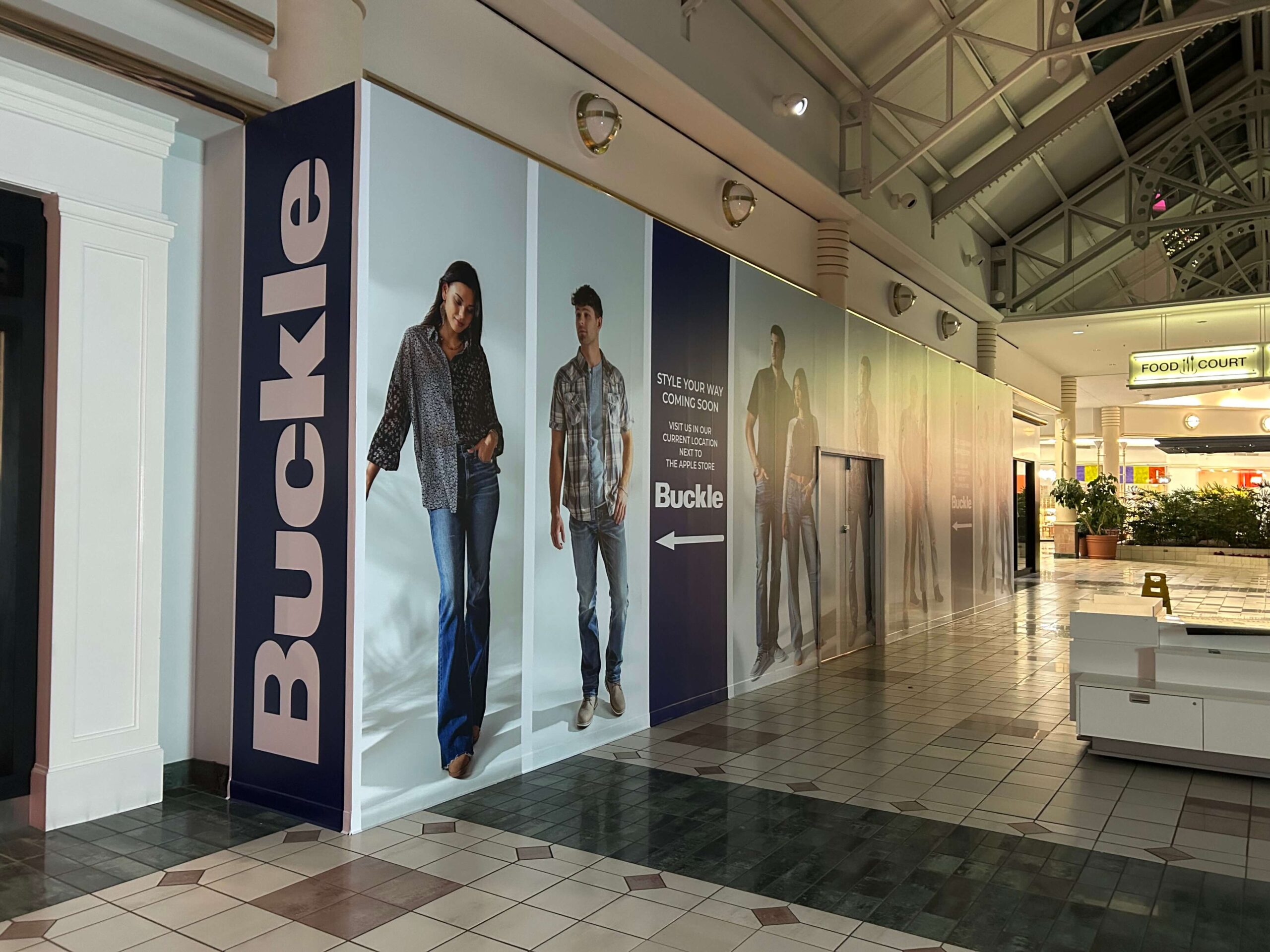 Full-height Buckle retail barricade installed in a shopping mall, featuring large lifestyle graphics of models, bold vertical Buckle branding, and directional messaging produced and installed by SuperGraphics.
