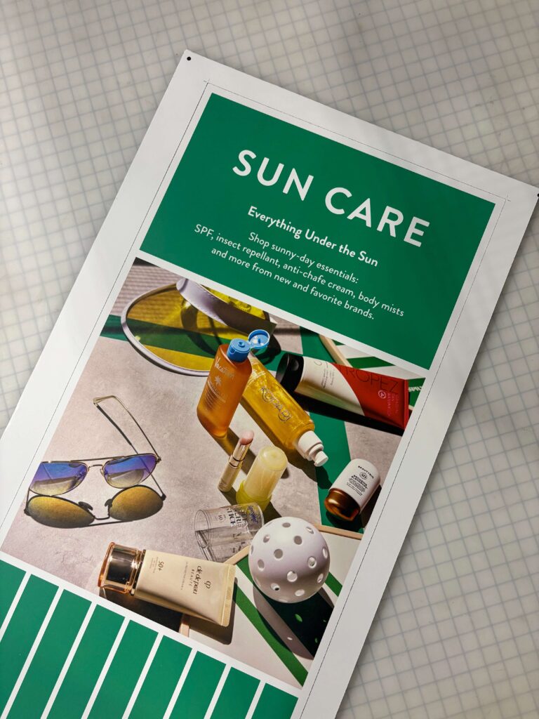 Printed sun care campaign sign produced by SuperGraphics for a major retailer, featuring green branding, summer product photography, and promotional messaging about SPF and warm-weather essentials.