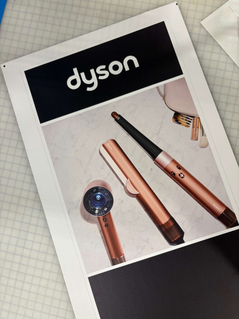 Printed display sign featuring Dyson hair-care tools, produced by SuperGraphics for a major retailer’s beauty department.