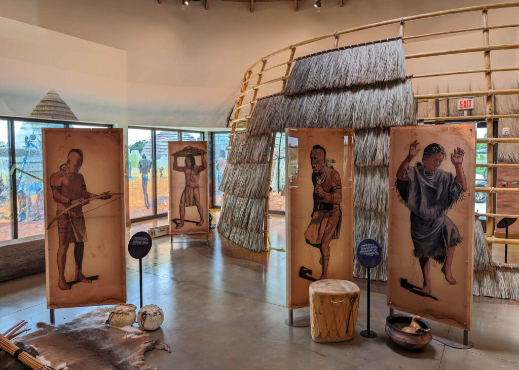 Museum exhibit featuring illustrated interpretive panels depicting Indigenous cultural practices, arranged around a thatched structure with artifact displays and immersive environmental elements, produced and installed by SuperGraphics.