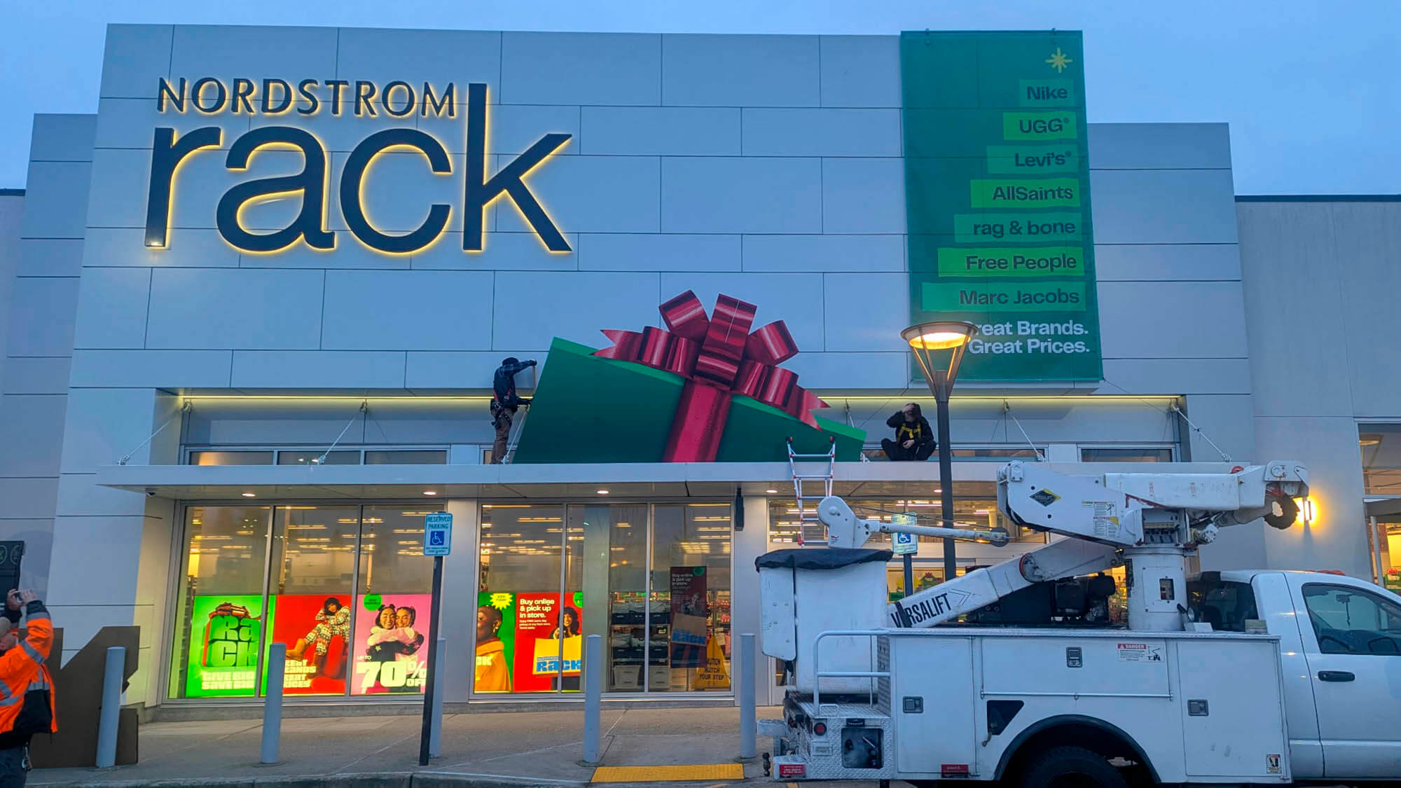 Nordstrom Rack storefront at dusk during installation of an oversized 3D holiday gift box display and large vertical banner for the 2025 Holiday campaign.