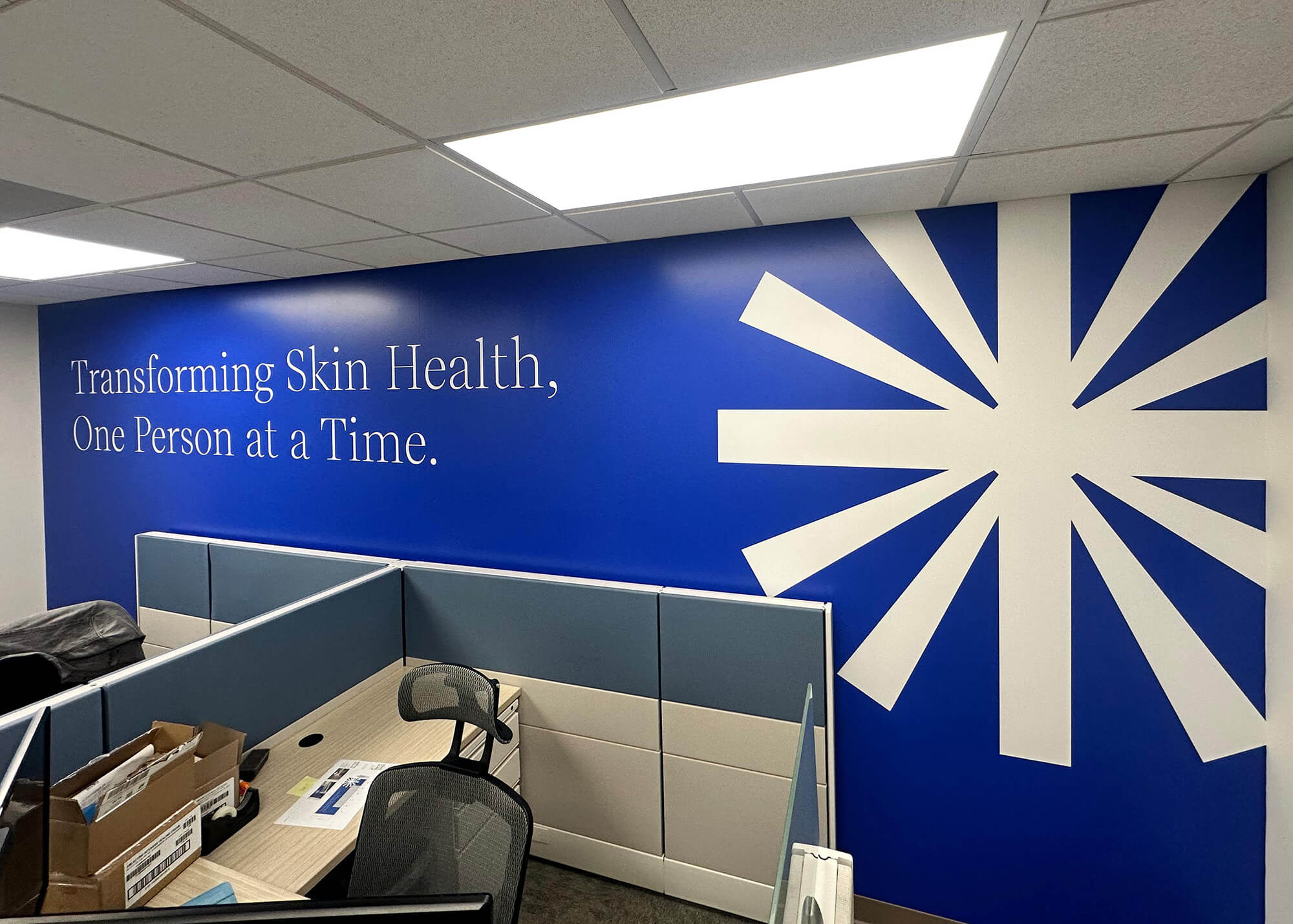 Advantice Health workspace wall mural featuring the tagline "Transforming Skin Health, One Person at a Time" in white text on a blue background with a large white starburst graphic, produced and installed by SuperGraphics.