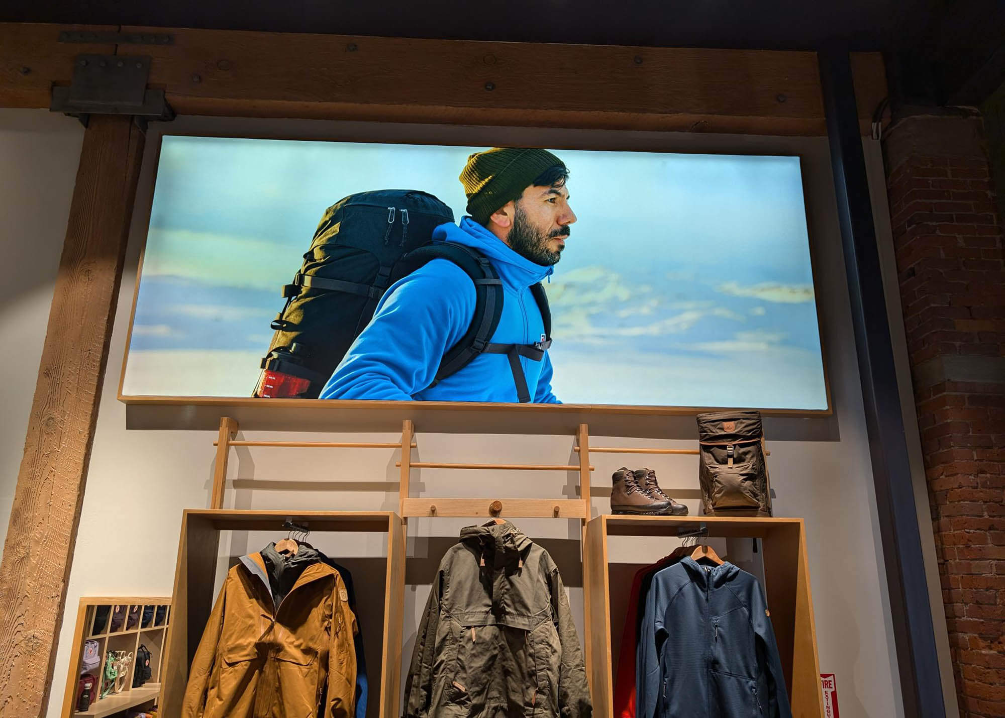 Fjallraven retail store backlit SEG fabric display of a hiker wearing a blue jacket and backpack, mounted above clothing racks and product shelving