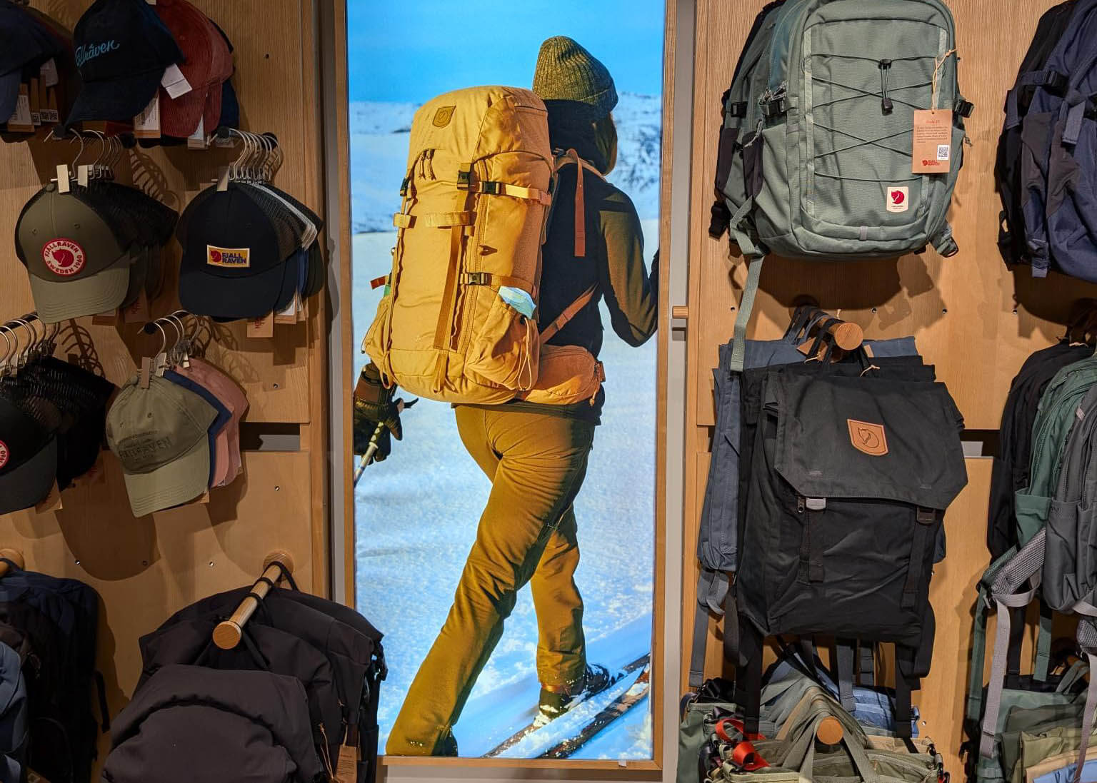 Fjallraven backlit SEG fabric display of a hiker with a yellow backpack trekking through snow, installed between retail shelving displaying hats and backpacks