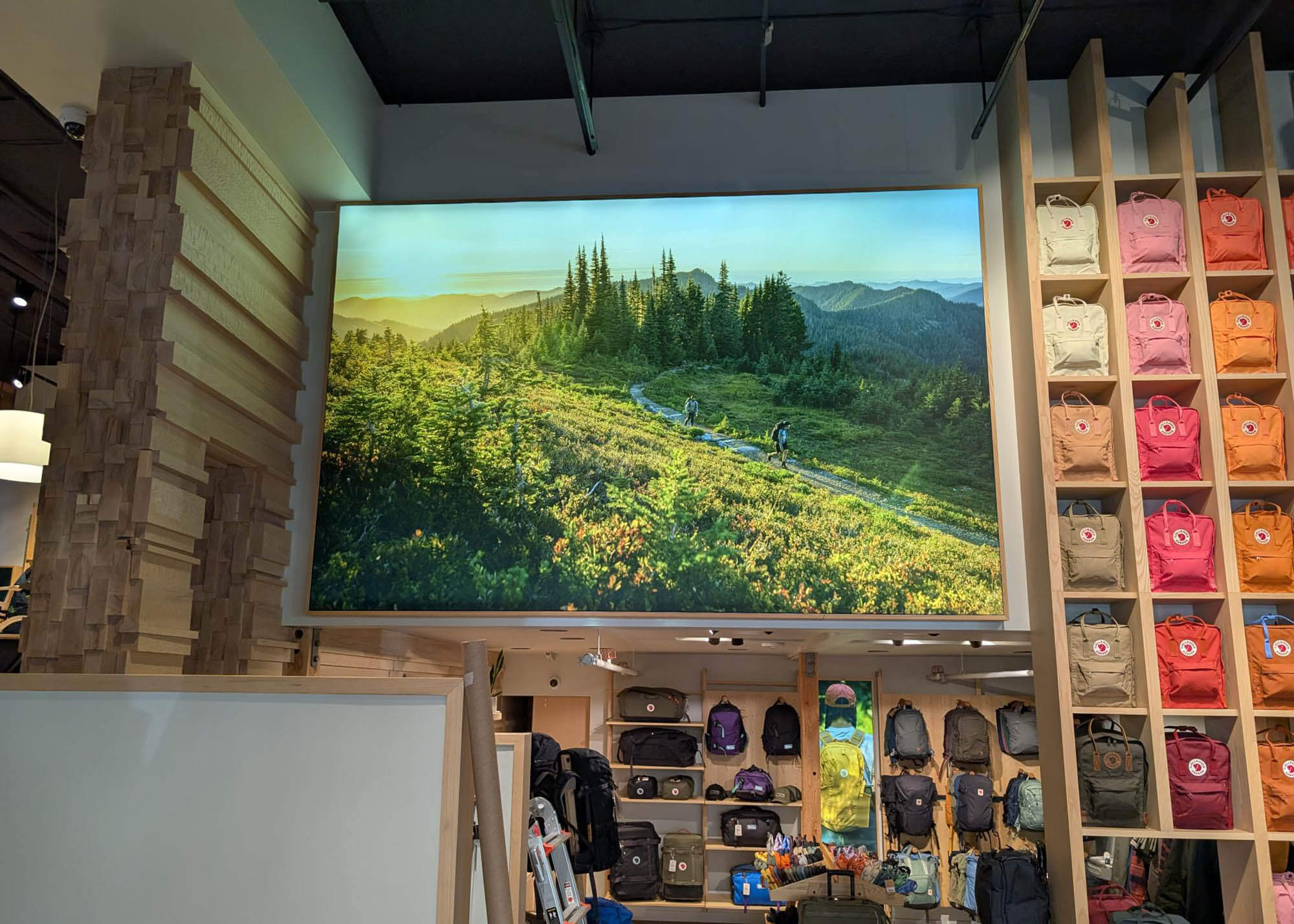 Large backlit Fjallraven SEG fabric display of hikers on a lush mountain trail at sunset, mounted above retail shelving with colorful Kanken backpacks and gear