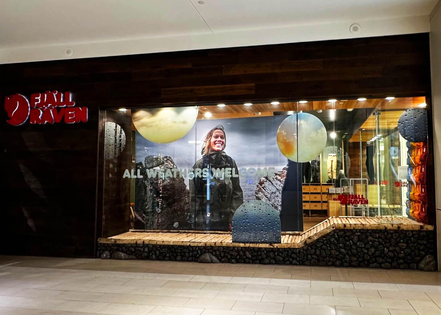 Fjallraven mall storefront with "All Weathers Welcome" window graphics featuring a smiling hiker in rain gear and circular rain droplet design elements, with illuminated Fjallraven logo above