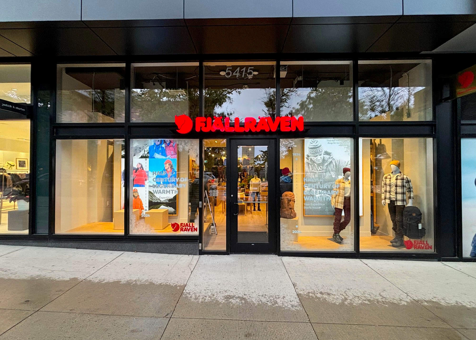 Fjallraven street-level storefront with illuminated red logo, "Half a Century of Legendary Warmth" campaign window graphics, and styled mannequins in the display windows