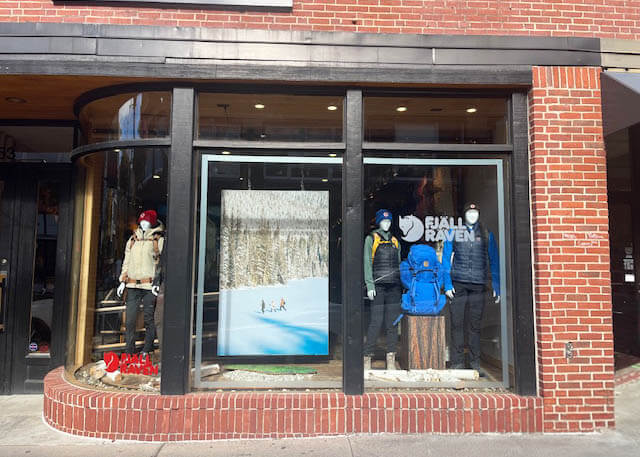 Fjallraven storefront with a center window graphic of a snowy winter landscape and cross-country skiers, flanked by dressed mannequins in outdoor apparel and backpacks, with the Fjallraven logo on the glass