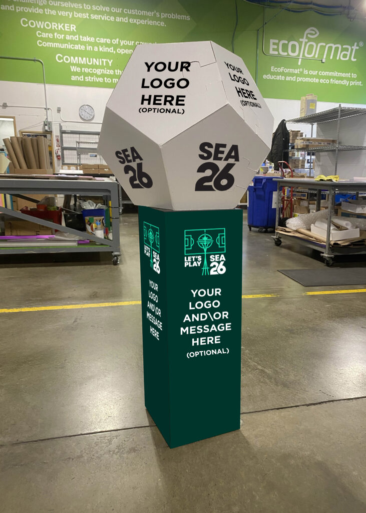 SuperGraphics oversized soccer ball display on a branded green pedestal with SEA 26 and Let's Play branding, shown in the SuperGraphics production facility.