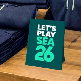 SuperGraphics SEA 26 retail kit table topper displaying "Let's Play SEA 26" in bold teal and white text, shown on a retail display surface.
