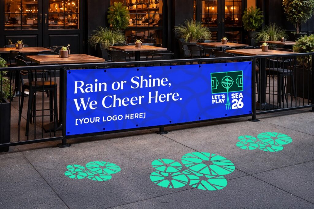 SuperGraphics branded watch party banner reading "Rain or Shine, We Cheer Here" displayed on an outdoor restaurant patio railing with soccer-themed floor decals, Seattle 2026 branding.