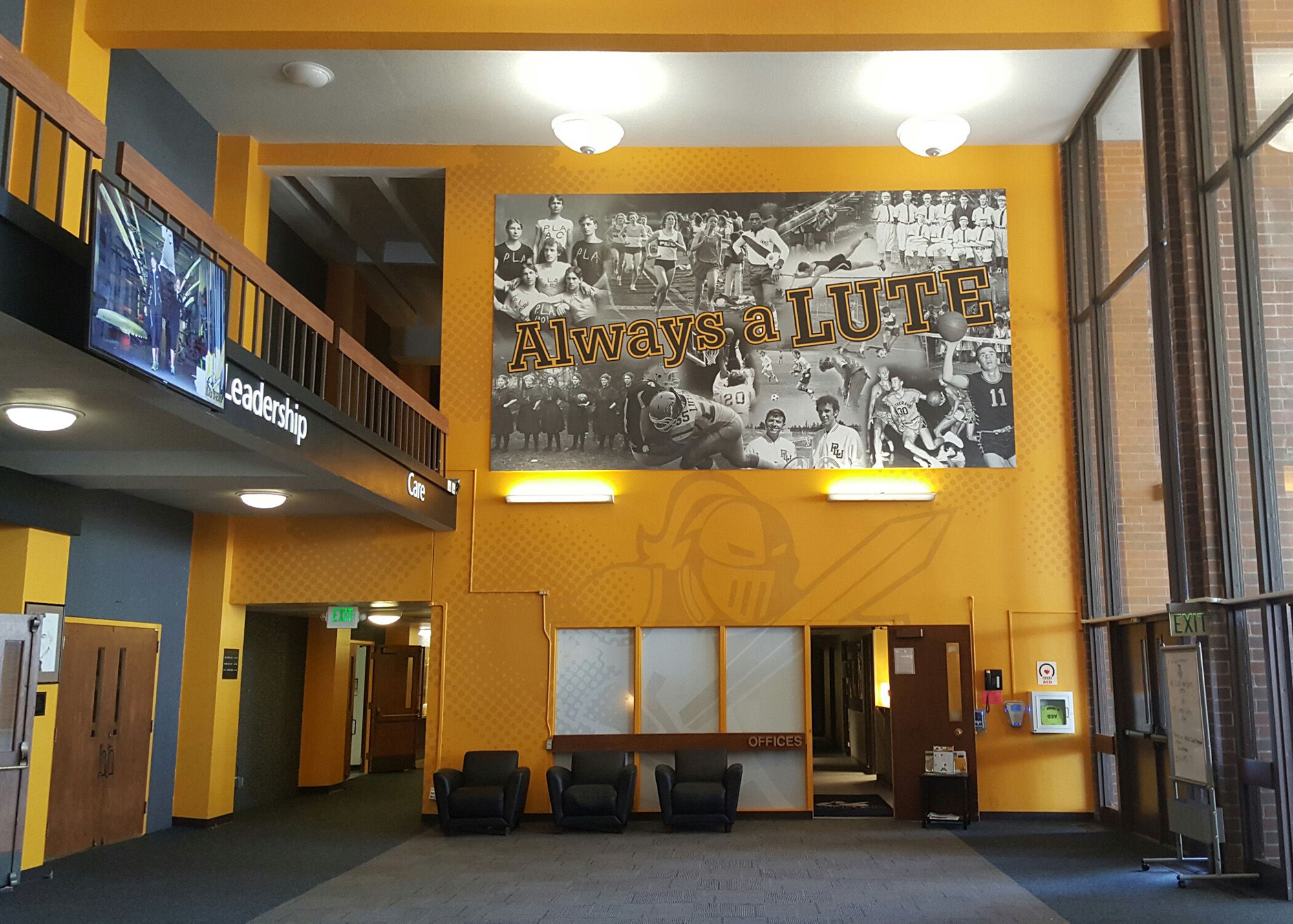 PLU athletics lobby featuring bold yellow walls with the Lutes mascot, a large "Always a LUTE" historical photo collage mural, and branded environmental graphics designed by SuperGraphics.