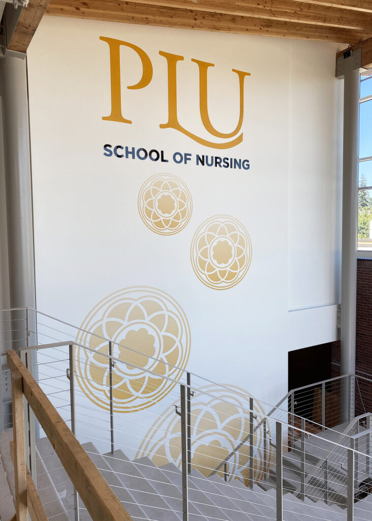 Large-scale interior wall mural for PLU School of Nursing featuring the university logo in gold and navy with repeating decorative medallion graphics, installed in a multi-story stairwell by SuperGraphics.
