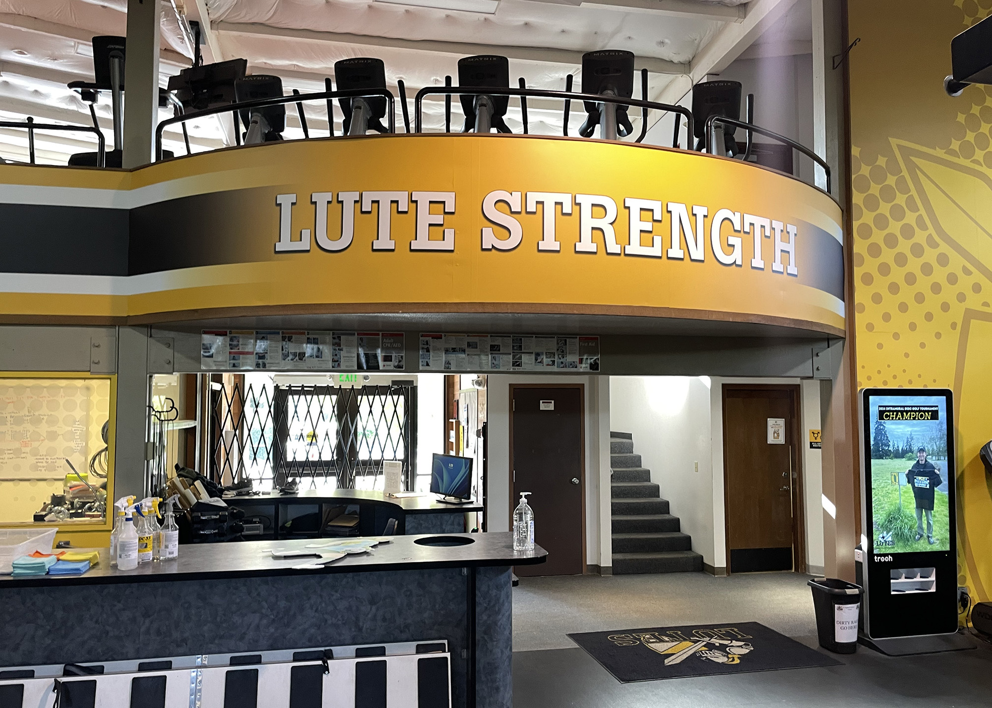 PLU's "Lute Strength" fitness center entrance featuring bold gold and black branded signage on a curved mezzanine, produced by SuperGraphics.