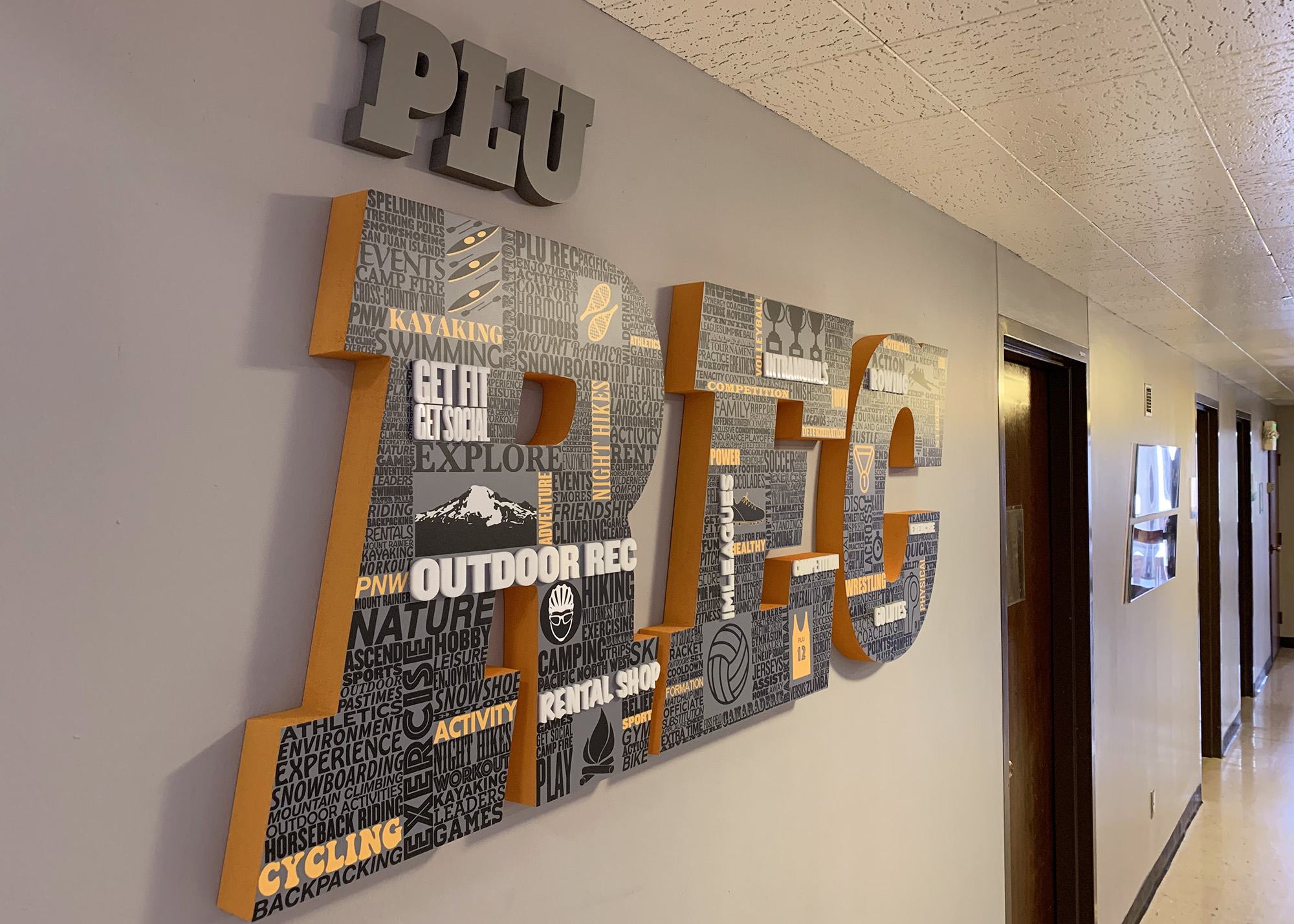 Large dimensional "PLU REC" lettering filled with activity-themed typography and icons mounted on a campus hallway wall, produced by SuperGraphics.