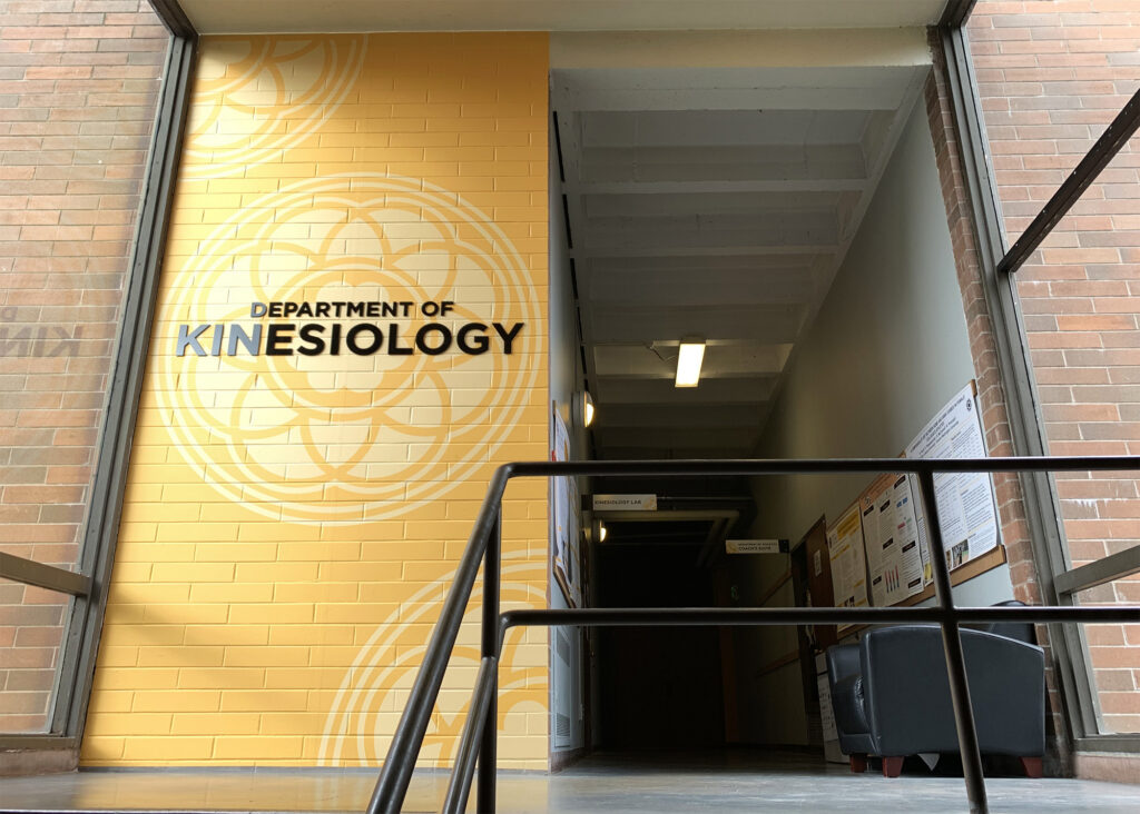PLU Department of Kinesiology entrance featuring a bold gold painted brick wall with the university's medallion graphic and dimensional "Department of Kinesiology" lettering, produced by SuperGraphics.