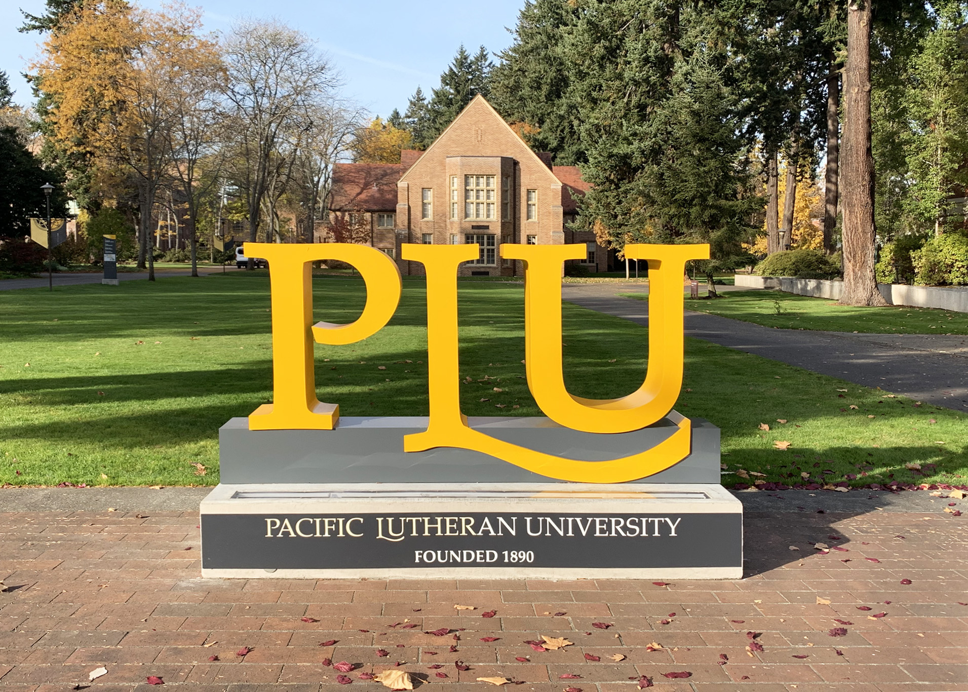 Large outdoor "PLU" dimensional campus sign reading "Pacific Lutheran University, Founded 1890" on a grey base, set against the university's brick buildings and autumn campus grounds.