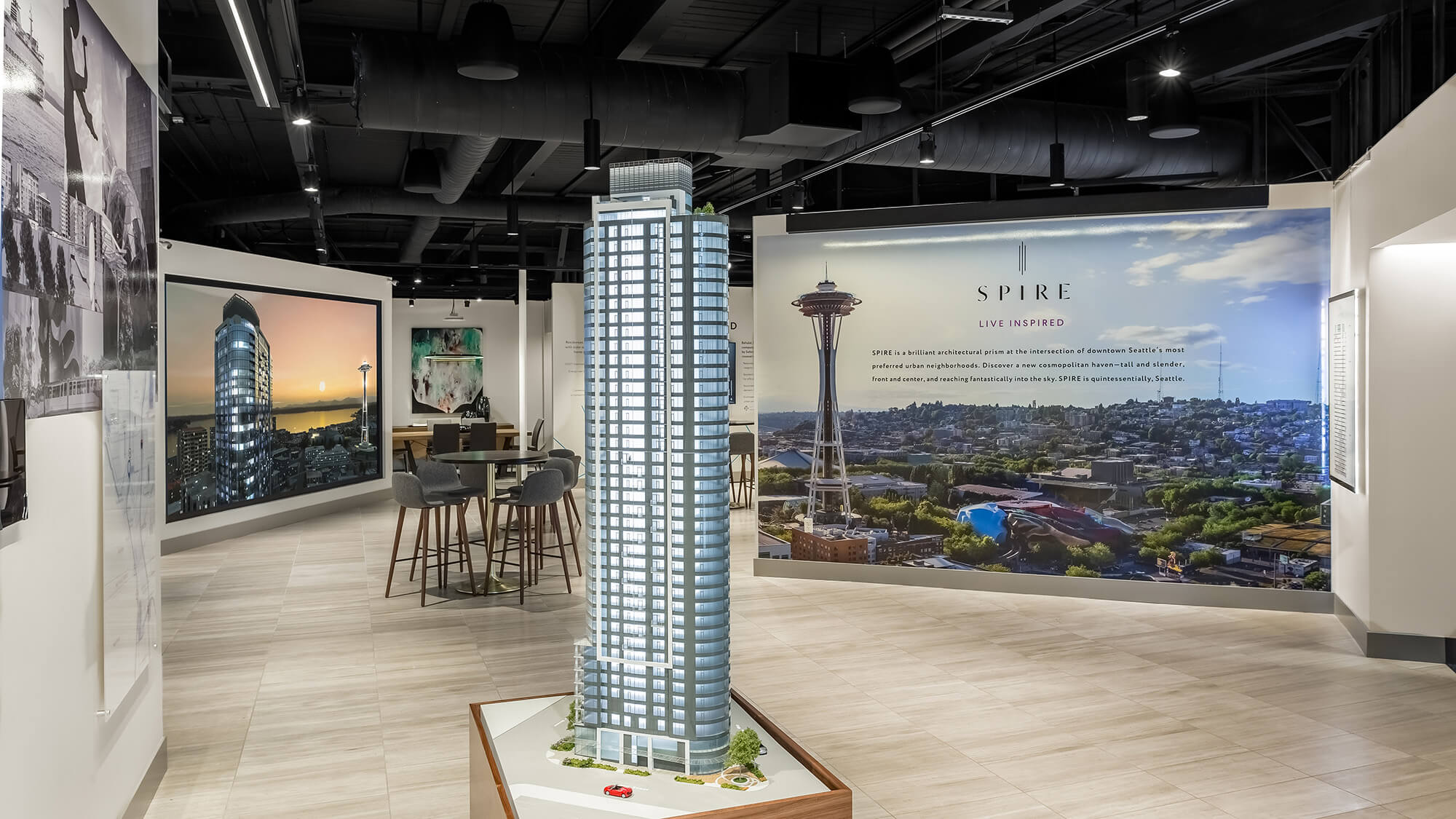 Interior of Sotheby's International Realty sales gallery featuring large-format wall graphics produced and installed by Supergraphics, including a dramatic aerial mural of Seattle's skyline with the Space Needle and Spire branding display, alongside an architectural scale model of the Spire high-rise tower.