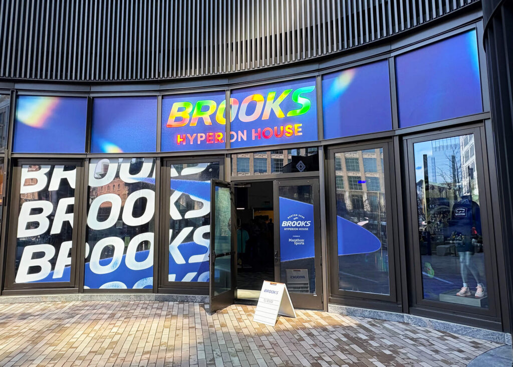 Storefront exterior with curved metal roof featuring colorful gradient signage reading "Brooks Hyperion House," large window graphics with blue and white geometric elements, and entry doors.