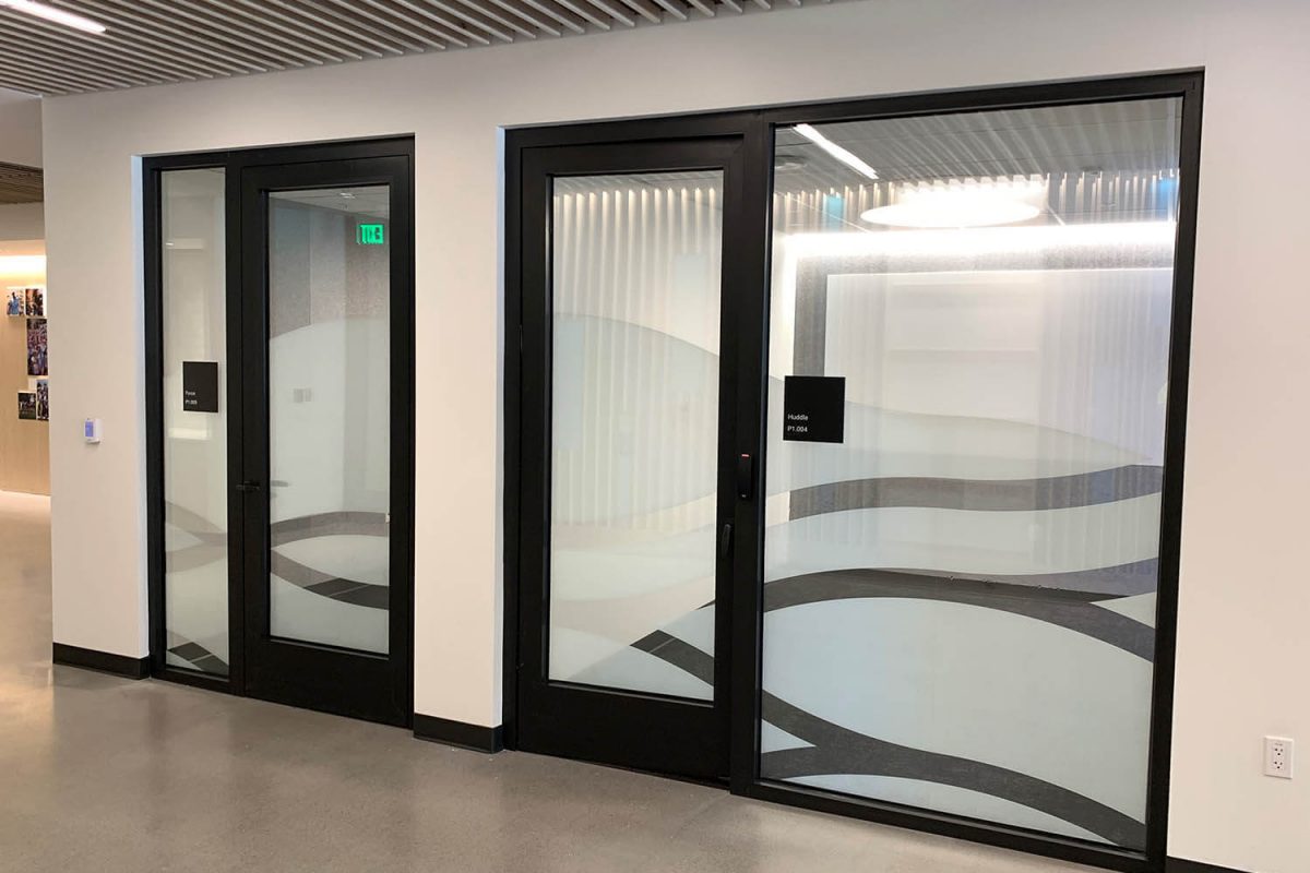 Frosted glass graphics with layered wave patterns applied to private office and huddle room storefronts in a modern workplace.