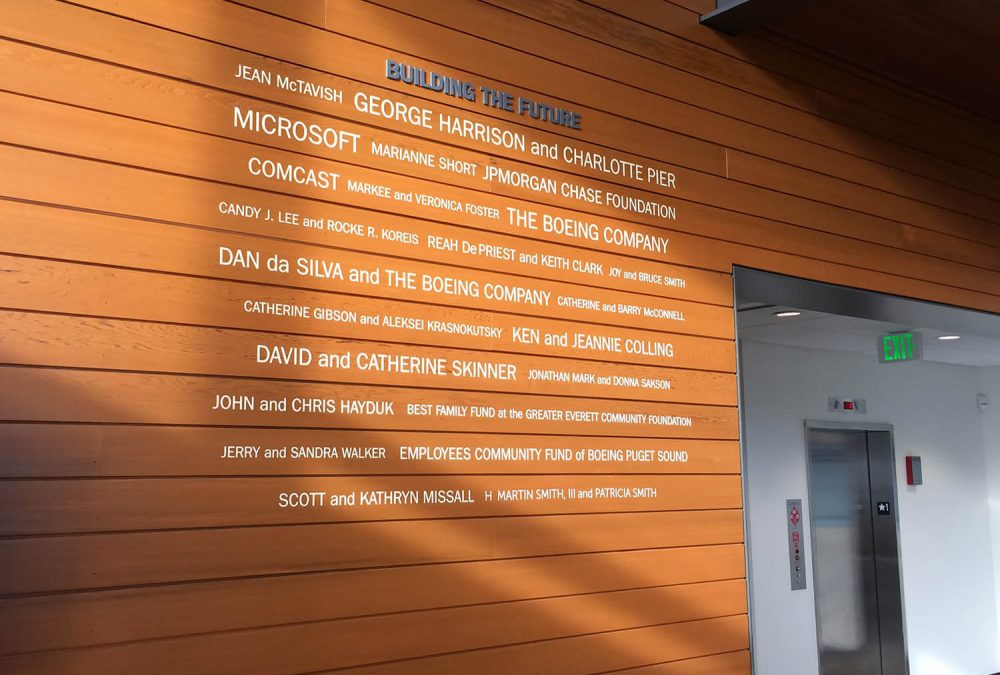 Wood-paneled donor recognition wall with raised metallic lettering displaying contributor names in a corporate lobby.