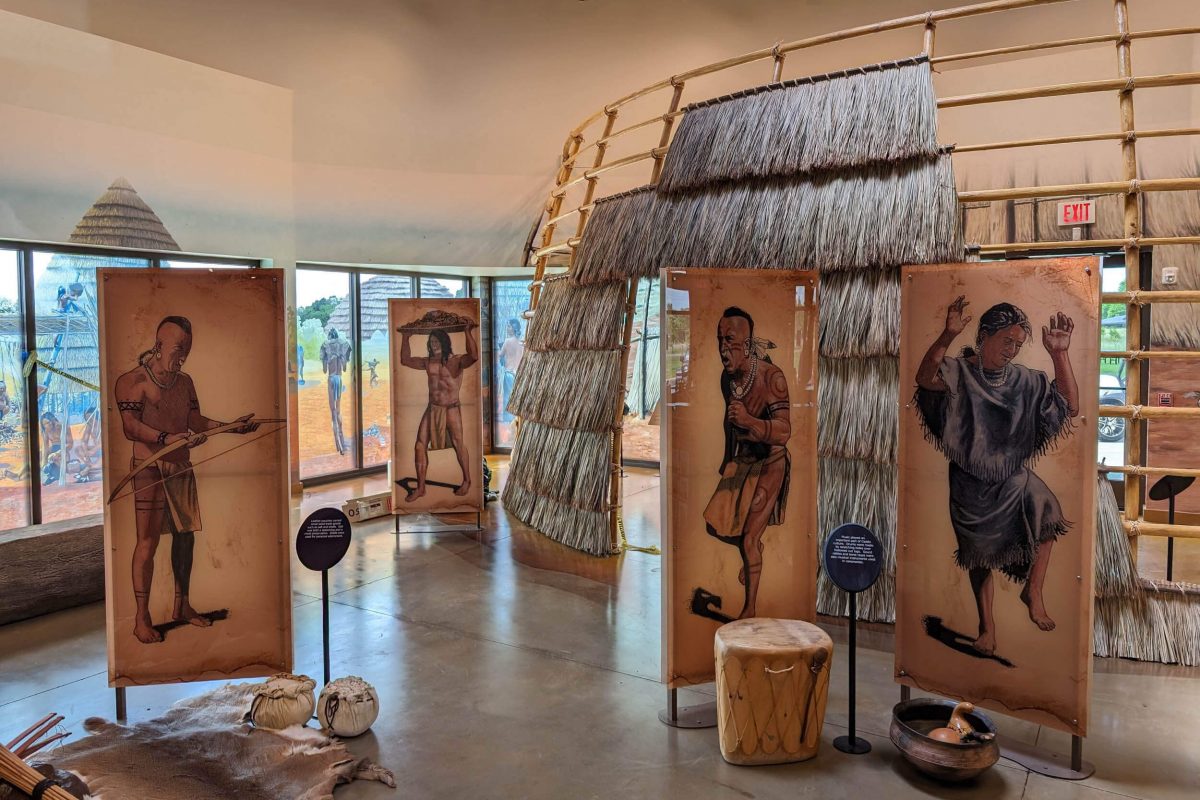 Museum exhibit featuring illustrated interpretive panels depicting Indigenous cultural practices, arranged around a thatched structure with artifact displays and immersive environmental elements, produced and installed by SuperGraphics.