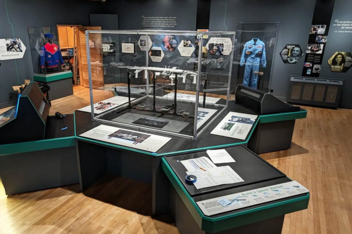 Interactive museum exhibit featuring central artifact display cases, angled interpretive panels, and educational graphics within a gallery environment, produced and installed by SuperGraphics.