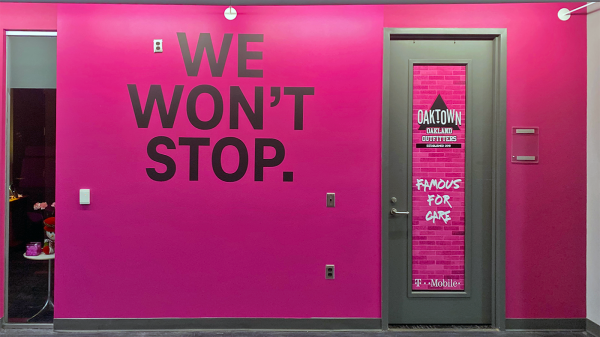 Wall and window graphic for T-Mobile, produced and installed by SuperGraphics. Showcases magenta T-Mobile branded graphics and "We Won't Stop" signage, as well as Oakland-specific branded window graphics on a glass door.