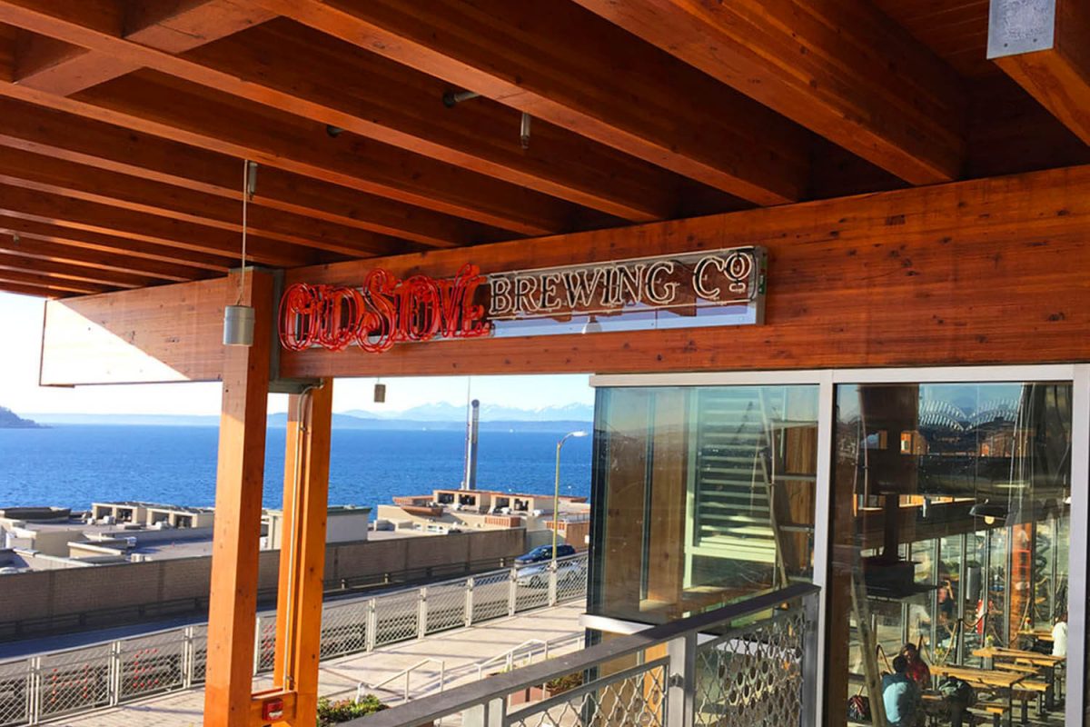 Exterior “Old Stove Brewing Co.” sign featuring red illuminated script lettering and rustic block lettering mounted on a wood beam overlooking the waterfront.