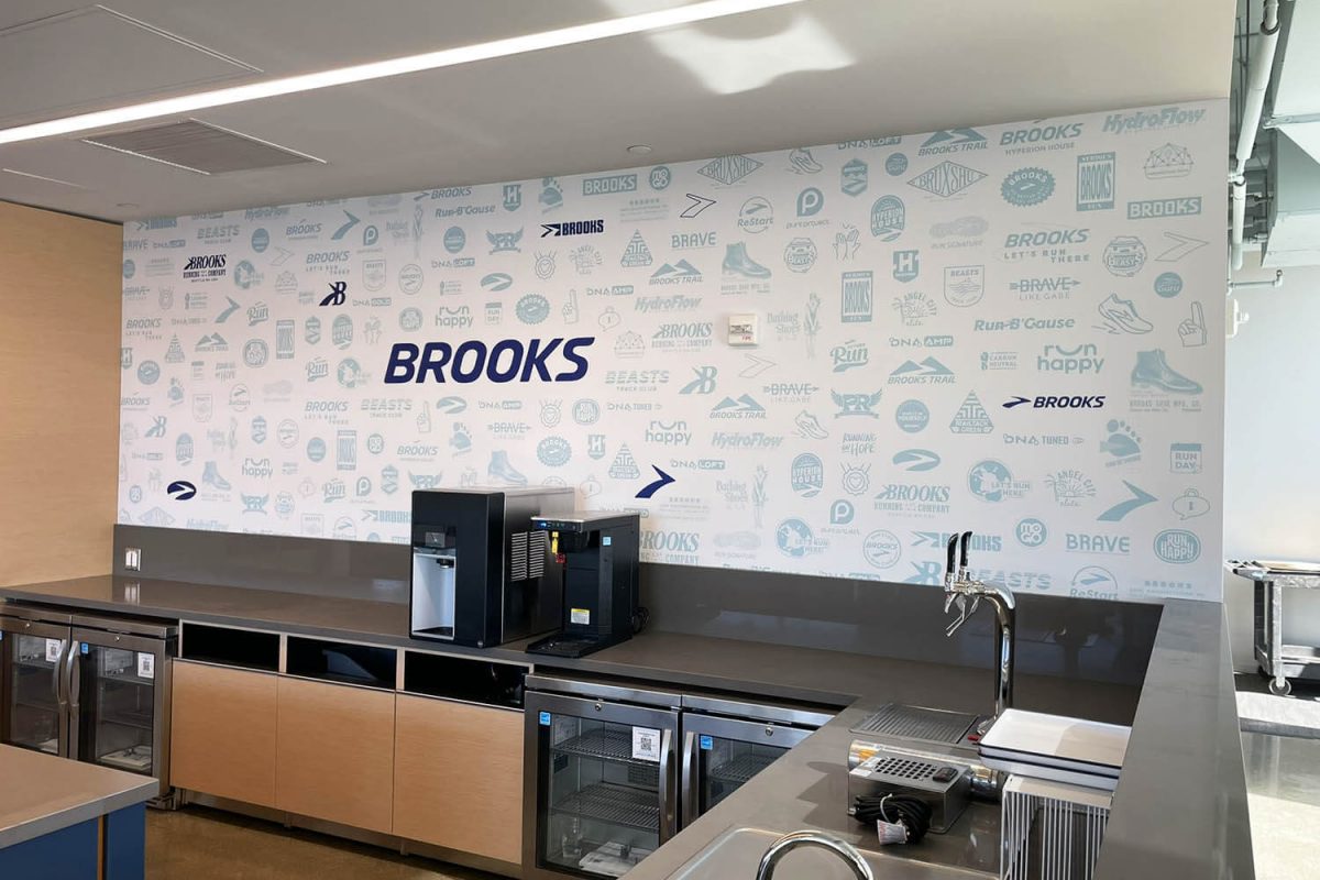 Brooks breakroom wall featuring a full-coverage pattern of Brooks logos, icons, and brand elements in blue tones with a large central Brooks wordmark.