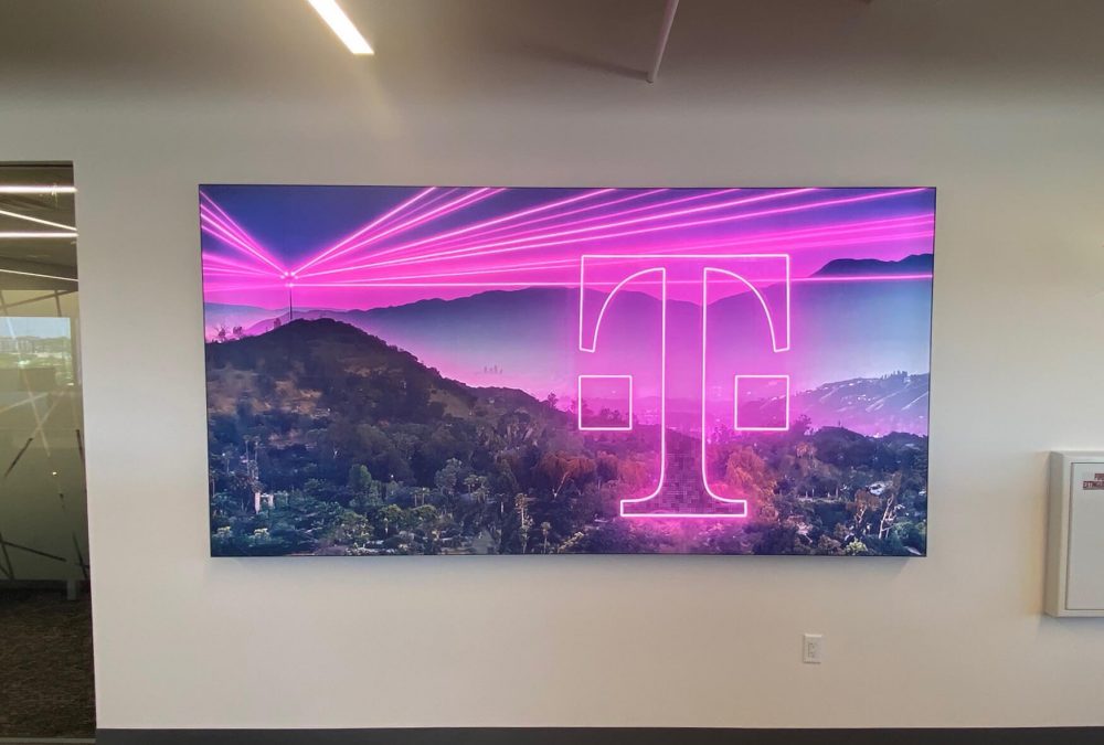 Large backlit SEG fabric frame featuring T-Mobile branding with magenta light rays over a hillside landscape installed in a corporate office by SuperGraphics
